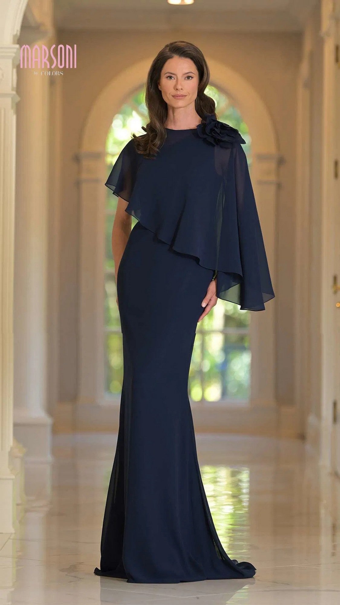 A woman stands indoors wearing the Marsoni MV1347, a long navy formal dress featuring chiffon fabric, an asymmetrical cape, and floral shoulder detail.